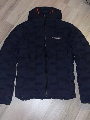 Dunjacka från Stellar - Snygg dunjacka från Stellar EQ med huva och dragkedja framtill. Jackan har diskreta orange detaljer och quiltad design som ger en modern look. Perfekt för kalla dagar och enkel att matcha med olika outfits.