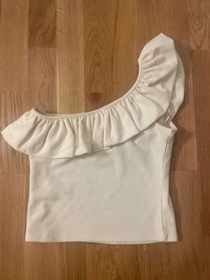 Vit oneshoulder topp med volang - Säljer en vit oneshoulder topp med bred volang längs halsringningen. Toppen är ärmlös och har en snygg asymmetrisk design som ger en trendig look. Perfekt för dig som vill sticka ut lite extra. Materialet känns mjukt och stretchigt.