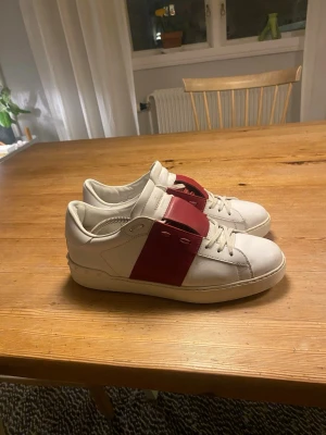 Valentino Open sneakers vit/röd - Snygga Valentino Open sneakers i vitt skinn med bred röd läderdetalj över snörningen. Klassisk låg modell med rund tå och låg sula. Skorna har små slitningar vid hälarna men märks inte när man har på sig de. Pris kan diskuteras. Storlek 41,5 men passar 42 perfekt.