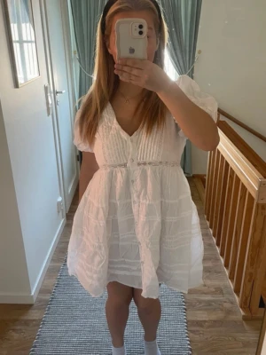 Vit kort klänning med puffärm - Söt vit kort klänning med puffärmar, v-ringning och knappar framtill. Klänningen har fina spetsdetaljer och lätt volang i kjolen. Perfekt för varma dagar och har en luftig passform.