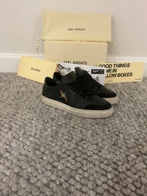 Svarta Axel Arigato sneakers med broderad fågel - Snygga svarta sneakers från Axel Arigato med broderad fågel i färg på sidan. Skorna har rund tå, snörning och vit platt sula. Tillverkade i skinn för en clean och modern look. Perfekta för dig som gillar stilrena detaljer och streetwear. Skorna är i ungefär skick 8 av 10. De är lite smutsiga men kan lätt tvättas bort. Tveka inte om ni har frågor. Priset kan diskuteras 😄🙌