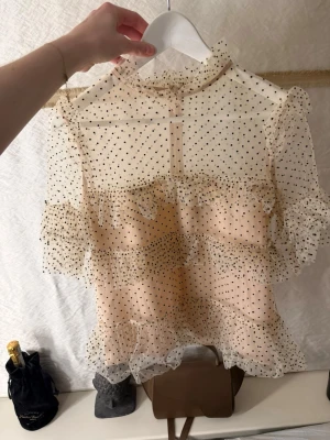 Prickig volangblus med mesh - Supersöt transparent blus i beige mesh med svarta prickar och volangdetaljer. Hög hals och korta ärmar, perfekt för lager på lager. Blusen har knäppning bak i nacken och är riktigt trendig med sitt romantiska uttryck.