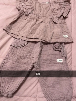 Ljuslila set från Newbie - Superfint tvådelat set från Newbie i ljuslila bomullsmuslin. Toppen har volangärmar och rynkad midja, byxorna har resår i midja och benslut samt dekorativa fickor. Perfekt för dig som gillar mjuka och luftiga material.