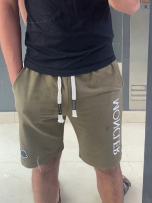 Olivgröna Moncler shorts med snörning - Snygga olivgröna shorts från Moncler med vit logga längs benet och snörning i midjan. Shortsen har två sidofickor och en liten Moncler-detalj på ena benet. Perfekta för sommaren och riktigt sköna att bära.