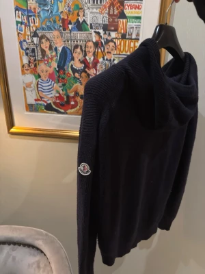 Blå stickad hoodie från Moncler i kashmir - Intresse koll på en blå Moncler Knit Cashmere hoodie från Moncler i mjuk blandning av Cashmere och Virgin Wool. En marinblå.  Tröjan har huva med dragsko och Moncler-logga på ärmen. Perfekt för dig som vill ha något både stilrent och bekvämt. SS22 Color Ink Blue Style / Ref num:  10919G00002M1900750