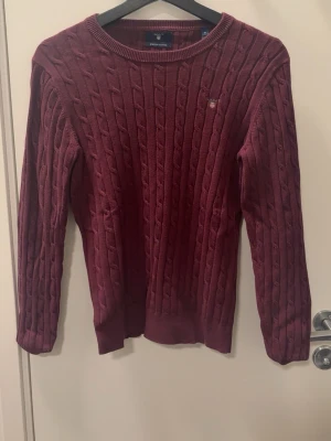 Vinröd kabelstickad tröja Gant - Vinröd kabelstickad tröja från Gant med rund halsringning och diskret logga på bröstet. Tröjan har långa ärmar och ribbade muddar vid ärmslut och nederkant. Perfekt för lager-på-lager och snygg till jeans.