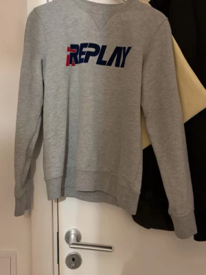 Grå sweatshirt från Replay - Snygg grå sweatshirt från Replay med tryckt logga i blått och rött på bröstet. Tröjan har rund halsringning, långa ärmar och ribbade muddar vid ärmslut och nederkant. Perfekt för en avslappnad och trendig stil.