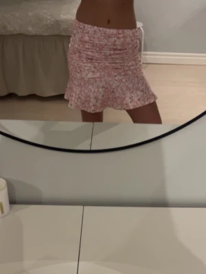 Rosa blommig volangkjol - Supersöt kort kjol i rosa med små vita blommor och volangkant. Kjolen har en figurnära passform upptill och är lätt utsvängd nedtill. Perfekt för dig som gillar en feminin och trendig stil.