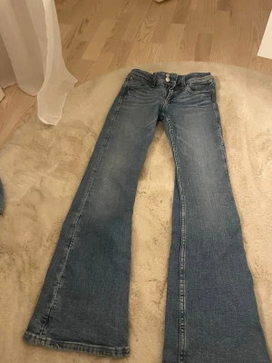 Blå bootcut jeans från Nudie Jeans - Säljer ett par klassiska blå bootcut jeans från Nudie Jeans med deras ikoniska broderade bakficka. Jeansen har normal passform och är tillverkade i slitstark bomull med snygg tvättad look. Perfekta för dig som gillar retrovibbar och vill ha ett par riktigt schyssta jeans.