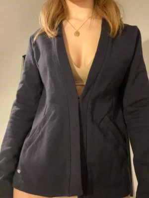 Mörkblå mjuk kavaj med fickor - Stilren mörkblå kavaj med djup v-ringning och två framfickor. Stängs där fram med hjälp av en liten krok och ögla. 😊