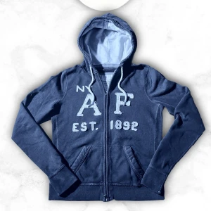 Mörkblå hoodie från Abercrombie & Fitch - Mörkblå hoodie med dragkedja från Abercrombie & Fitch. Stor logga och text 'NY A&F EST. 1892' framtill, två fickor och justerbar huva med vita snören. Mjuk insida och klassisk collegekänsla, perfekt för en avslappnad stil. Passar någon 160-170