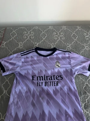 Lila Real Madrid matchtröja - Säljer en lila Real Madrid matchtröja  med svart krage och muddar. Tröjan har ett geometriskt mönster, klubbmärke på bröstet och 'Emirates Fly Better' tryckt framtill. Perfekt för fotbollsträning eller att bära till vardags.
