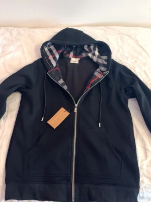 Svart hoodie från Burberry med dragkedja - Svart hoodie från Burberry med hel dragkedja och klassiskt rutigt foder i huvan. Den är helt oanvänt säljer den pga av att den passar inte mig storleken är L men möjligtvis passa M också om du är osäker skriv läng och vikt. Den har inga problem helt ny.