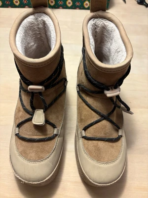 Beige boots från Tretorn - Snygga beige boots från Tretorn (Aspa Hybrid Low) med mjukt foder och detaljer i mocka och syntet. Skorna har svarta snören med reflexdetaljer och praktiska dragflikar bak. Perfekta för kalla dagar när du vill hålla stilen.
