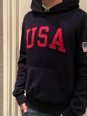 Mörkblå hoodie USA Polo Ralph Lauren - Mörkblå hoodie från Polo Ralph Lauren med stor röd 'USA'-text på bröstet och amerikansk flagga på ärmen. Klassisk känguruficka framtill och huva med dragsko. Perfekt för dig som gillar sportig och avslappnad stil.