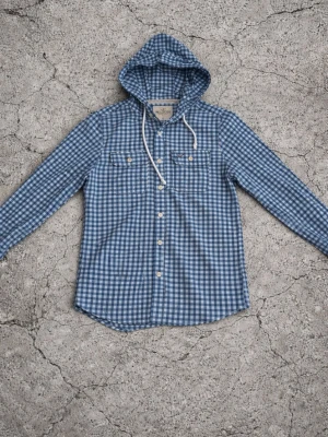 Blårutig hoodie från Hollister - Snygg blårutig hoodie från Hollister med vita knappar och två bröstfickor. Klassisk huva med vita snören och det ikoniska röda Hollister-loggan broderad på ena fickan. Tillverkad i mjukt bomullstyg som känns skönt mot huden.