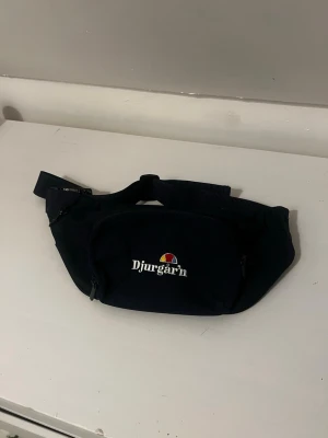 Blå axelväska Djurgår'n - Snygg blå axelväska från Djurgår'n med justerbart band och dragkedja. Väskan har ett broderat Djurgår'n-tryck framtill och en praktisk framficka. Perfekt för att bära det viktigaste när du är på språng.