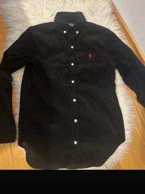 Svart manchester-skjorta Polo Ralph Lauren - Snygg svart manchester-skjorta från Polo Ralph Lauren med klassisk button-down krage och vita knappar. På bröstet finns den ikoniska röda logotypen. Skjortan har lång ärm och är tillverkad i mjuk manchester för en stilren och bekväm look.