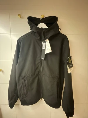 Soft Shell jacka från Stone Island - Snygg svart vindjacka från Stone Island med huva och dragkedja framtill. Jackan har två stora fickor, ribbade muddar och den klassiska logotypen på ärmen. Perfekt för dig som vill ha en stilren och funktionell jacka.