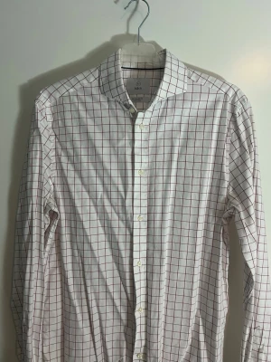 Vit skjorta med rött rutmönster från Bläck - Klassisk vit skjorta från Bläck med tunna röda rutor. Skjortan har lång ärm, knäppning framtill och en smal krage. Tillverkad i ett lätt bomullstyg som känns skönt mot huden. Perfekt för dig som gillar stilrena och tidlösa plagg.