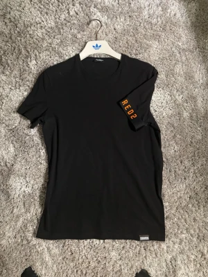 Svart t-shirt från Dsquared2 - Svart t-shirt från Dsquared2 med orange texten 'RED2' tryckt på ena ärmen. Klassisk rund halsringning och normal passform. Tillverkad i mjuk bomull som känns skön mot huden. Perfekt för en clean och stilren look.