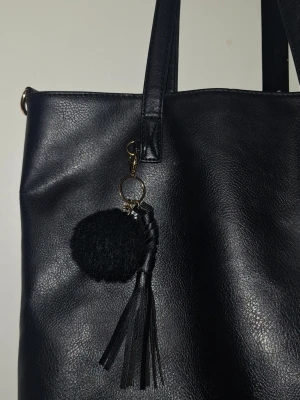 Handväska - Sthlm stil stilren svart handväska i fejkläder med långa axelband. Väskan har en dekorativ nyckelring med en fluffig boll och svarta lädertofsar. Perfekt för dig som gillar enkel men trendig design. Rymlig modell som rymmer det mesta du behöver. Passar som t ex. en skolväska, när man ska resa, weekend bag etc.