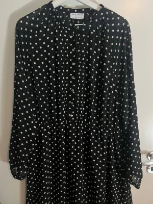 Svart prickig klänning Holly & White - Svart långärmad klänning från Holly & White med vita prickar och knappar framtill. Klänningen har en avslappnad passform och lätt rynkad midja. Perfekt för dig som gillar klassiska mönster och enkel stil.