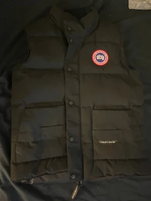 Canada goose väst - Snygg svart dunväst från Canada Goose med klassisk logga på bröstet och stora fickor framtill. Västen har hög krage, tryckknappar och dragkedja. Perfekt för lager-på-lager och håller dig varm under kyliga dagar. (Möts upp bara Helsingborg)