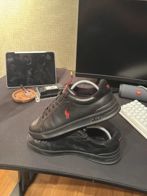 Svarta Polo Ralph Lauren sneakers - Snygga svarta sneakers från Polo Ralph Lauren med klassisk röd logga på sidan. Skorna har rund tå, platt sula och snörning. Tillverkade i skinn med diskreta detaljer och låg profil, perfekta för en clean och stilren look. 