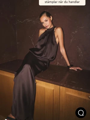 Brun halterneck långklänning i satin - Snygg brun långklänning i satin med halterneck och lös passform. Klänningen har ärmlös design och ett elegant, glansigt material som ger en lyxig känsla. Perfekt för dig som vill sticka ut med en stilren och modern look. Med prislapp!!