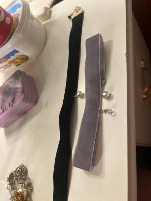 Två choker-halsband i sammet - Säljer två trendiga choker-halsband, ett i svart sammet med guldfärgat spänne och ett i ljusgrå sammet med silverfärgade detaljer och små berlocker. Perfekta accessoarer för att lyfta din outfit med en cool och stilren touch.