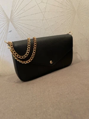 Svart kuvertväska från DonDonna - Stilren svart kuvertväska från DonDonna med guldfärgad kedja och enkel knappstängning. Väskan har en slät, elegant yta och klassisk rektangulär form som passar perfekt till alla tillfällen där du vill ha en clean look.