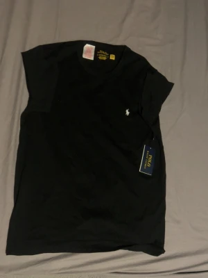 Svart t-shirt från Polo Ralph Lauren - Klassisk svart t-shirt från Polo Ralph Lauren med den ikoniska lilla logon broderad på bröstet. T-shirten har rund hals och korta ärmar. Tillverkad i mjuk bomull som känns skön mot huden. Perfekt basplagg för en clean och stilren look. Org pris 650kr