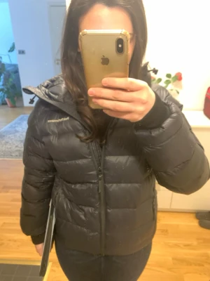 Svart dunjacka Peak Performance - Snygg svart dunjacka från Peak Performance med huva och dragkedja framtill. Jackan har en quiltad design, två sidofickor med dragkedja och diskret logga på bröstet. Perfekt för kalla dagar och riktigt skön passform.