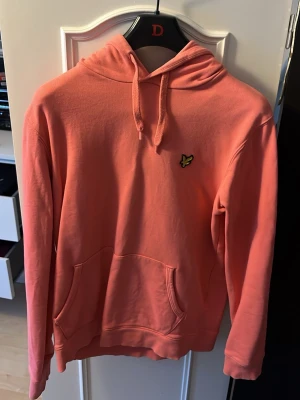 Korallfärgad hoodie från Lyle & Scott - Säljer en snygg korallfärgad hoodie från Lyle & Scott med klassisk gul logga på bröstet. Hoodien har huva med dragsko och en stor magficka framtill. Tillverkad i mjukt bomullsmaterial och har ribbade muddar vid ärmar och nederkant.