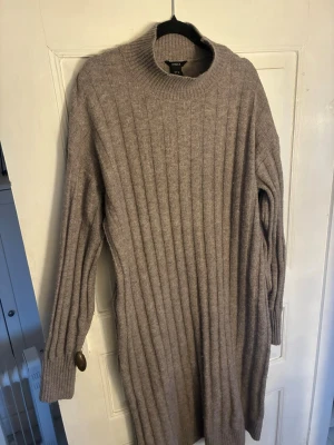 Beige/grå ribbad stickad klänning Lindex - Mysig beige/grå stickad klänning från Lindex med ribbad struktur och lång ärm. Perfekt för kyligare dagar och enkel att styla med boots eller sneakers. Rund halsringning och mjukt material som känns skönt mot huden.