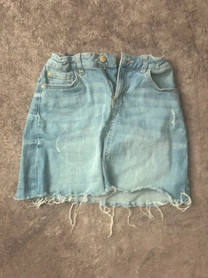 Ljusblå jeanskjol med fransig kant - Trendig ljusblå jeanskjol med rå, fransig nederkant och klassiska fem fickor. Kjolen har knapp och dragkedja framtill samt bälteshällor. Perfekt för en avslappnad och cool stil.