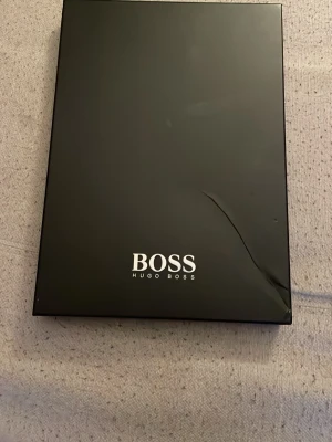 Svart axelväska från Hugo Boss - Stilren svart axelväska från Hugo Boss med stort BOSS-tryck framtill. Väskan har en justerbar axelrem och en praktisk dragkedja på framsidan. Perfekt för att bära dina viktigaste saker med stil.