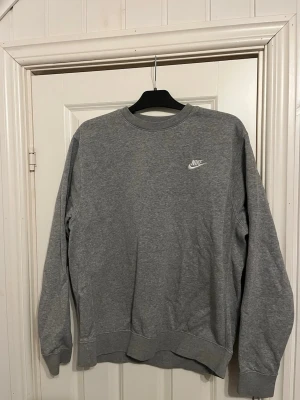 Grå sweatshirt från Nike - Klassisk grå sweatshirt från Nike med rund halsringning och diskret vitt Nike-logga på bröstet. Tröjan har långa ärmar och ribbade muddar vid ärmslut och nederkant. Perfekt för en avslappnad och sportig stil.