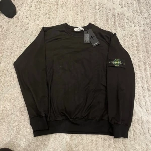 Svart sweatshirt från Stone Island - Svart sweatshirt från Stone Island med klassisk rund hals och ikonisk logopatch på vänster ärm. Tröjan har ribbade muddar vid ärmslut och nederkant. Perfekt för en avslappnad och stilren look.