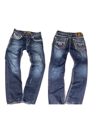 Blåa baggy/relaxed jeans (Rock Revival stil) - Snygga blå jeans (Rock Revival ish) med tydliga vita kontrastsömmar och slitningar(menade till att vara där). Jeansen har raka ben, klassiska fem fickor och detaljerade bakfickor med broderi. Perfekta för dig som vill sticka ut med din denimstil. DM för fler eventuella frågor!😇