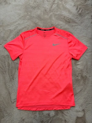 Nike Running Miler - Snygg neonrosa löpartröja från Nike med reflekterande logga på bröstet och diskreta grå detaljer vid axlarna. Tillverkad i lätt och snabbtorkande funktionsmaterial som andas, perfekt för löpning och träning.