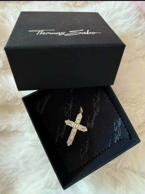 Thomas Sabo korshalsband i silver - Säljer ett snyggt korshalsband från Thomas Sabo i silverfärg med gnistrande stenar. Hänget har en klassisk korsform och är dekorerat med rektangulära och runda klara stenar som ger ett lyxigt intryck. Perfekt accessoar för dig som gillar statement-smycken.