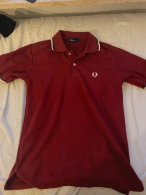 Vinröd pikétröja från Fred Perry - Säljer en vinröd pikétröja från Fred Perry med vit logga på bröstet och vita detaljer på kragen. Klassisk polokrage och korta ärmar. Perfekt för dig som gillar stilrena och sportiga plagg.