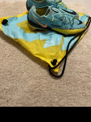 Nike Phantom fotbollsskor - Säljer ett par Nike Phantom  fotbollsskor i ljusblått med svarta och gula detaljer. Skorna har snörning och är tillverkade i syntetmaterial för bästa grepp och kontroll på planen. Swoosh-logga på sidan och cool design som sticker ut. Bara använt skorna 1 gång. 