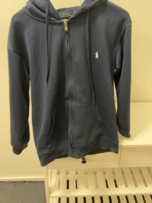 Marinblå hoodie från Polo Ralph Lauren - Mörkblå hoodie med dragkedja från Polo Ralph Lauren. Klassisk design med vit logga på bröstet, huva med dragsko och två fickor framtill. Perfekt för en chill och stilren look.