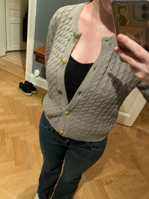 Grå kabelstickad kofta från H&M - Grå kabelstickad kofta från H&M med guldiga knappar framtill. Koftan har rund halsringning och ribbade muddar vid ärmslut och nederkant. Perfekt att slänga över en topp för en snygg lager-på-lager-look.