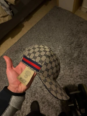 Gucci keps med GG-mönster - Snygg keps från Gucci i beige med klassiskt GG-monogram över hela kepsen. Har en röd och marinblå rand baktill som signaturdetalj. Tillverkad i canvasmaterial med böjd skärm och justerbar rem bak. Äkta med kvitto och paket till