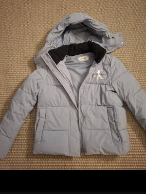 Ljusblå pufferjacka från Calvin Klein - Snygg ljusblå pufferjacka från Calvin Klein Jeans med huva och dragkedja. Jackan har ett stort CK-logga tryckt på bröstet, perfekt för kyliga dagar. Nästan oanvänd