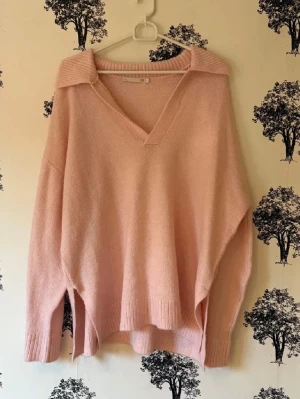 Ljusrosa stickad tröja med krage - Oversized stickad tröja i ljusrosa med djup V-ringning och bred krage. Tröjan har långa ärmar, ribbade muddar och slitsar i sidorna. Perfekt för lager-på-lager och en chill look.